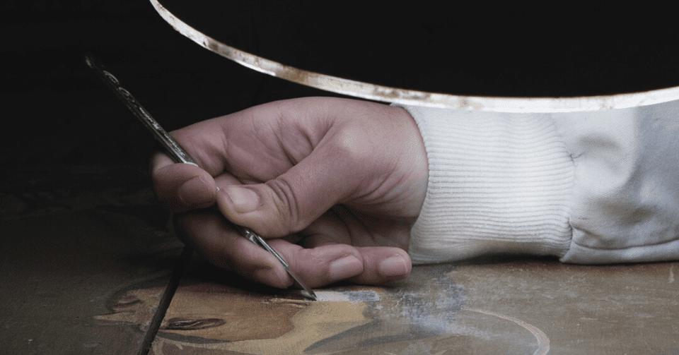 A imagem retrata um momento de intervenção técnica sobre uma obra de arte sacra, evidenciando o cuidado e a precisão que definem o trabalho da Dalmática. Sob iluminação controlada, um restaurador aplica técnicas especializadas para preservar os detalhes originais de uma pintura histórica. Este processo reflete o compromisso da empresa com a autenticidade, a ética e a valorização do património religioso.