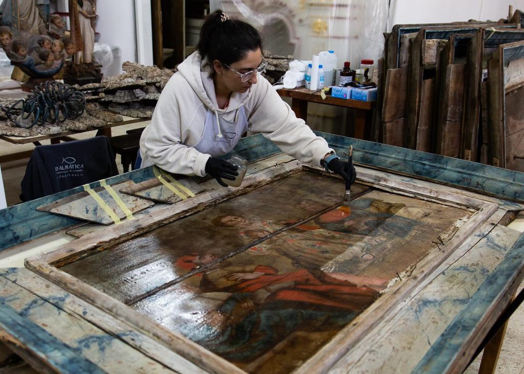 Nesta imagem, captamos um momento íntimo do processo de conservação de uma obra de arte histórica. Um dos nossos restauradores dedica-se meticulosamente à recuperação de uma pintura religiosa, utilizando técnicas especializadas e materiais adequados para preservar a integridade original da peça. Equipado com proteção e rodeado por ferramentas e obras em diferentes fases de intervenção, este profissional representa o rigor e a sensibilidade que definem o trabalho da Dalmática. Cada gesto é guiado pelo compromisso de devolver ao património a sua dignidade estética e espiritual, respeitando a memória que carrega.
